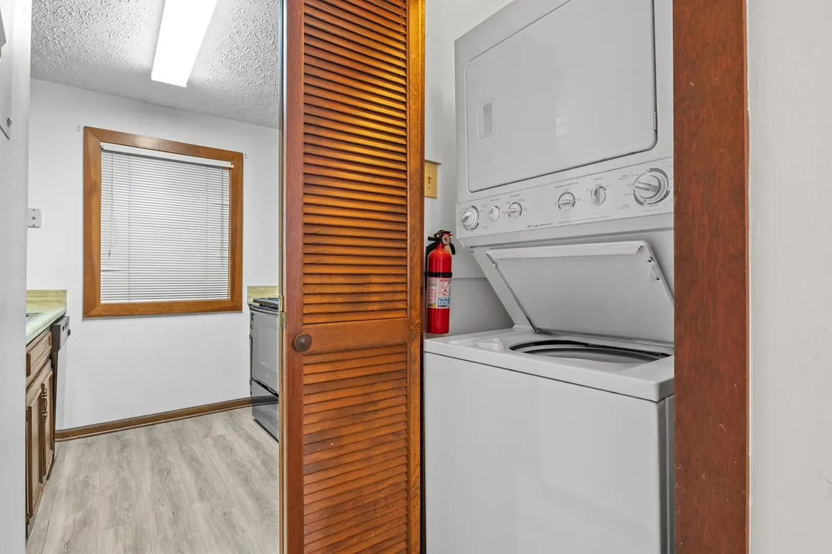A laundry nook featuring a full-size washer and dryer, neatly tucked behind wooden shutter doors. The space is practical and efficient, with a window providing natural light and a fire extinguisher for safety.