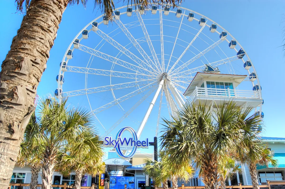 The Myrtle Beach SkyWheel stands tall against a festive backdrop of palm trees and the entrance plaza. The vibrant scene suggests excitement and family adventure, showcasing one of the local attractions included in the Free Fun Program.