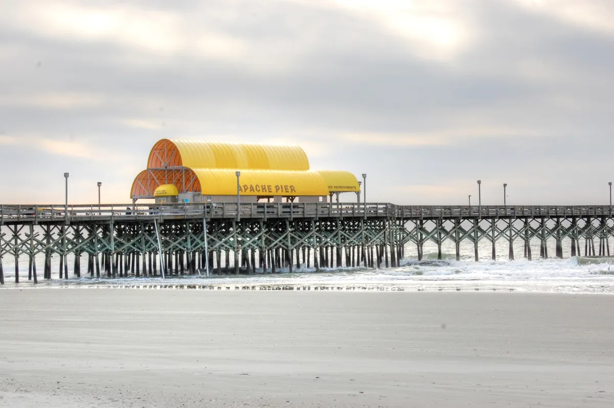 This image captures the iconic Apache Pier, with its distinctive yellow-roofed building and sturdy wooden structure extending out into the ocean. The serene seascape and gentle colors evoke a sense of relaxation and adventure, inviting families to visit the pier for fishing, strolling, or enjoying sunrise views.