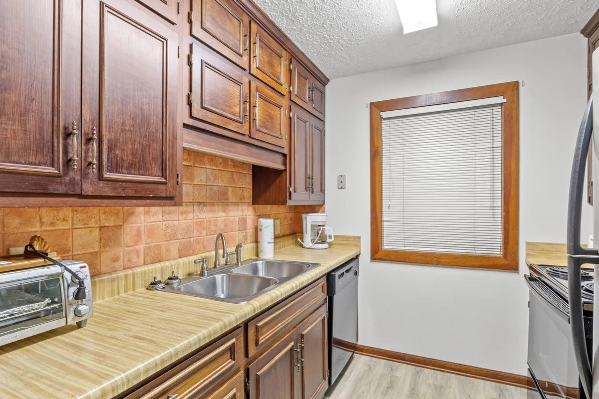 A cozy kitchen view highlighting wooden cabinetry, a kitchen sink, and a window with blinds. The space feels intimate and functional, ideal for everyday routines like morning coffee or baking with family.