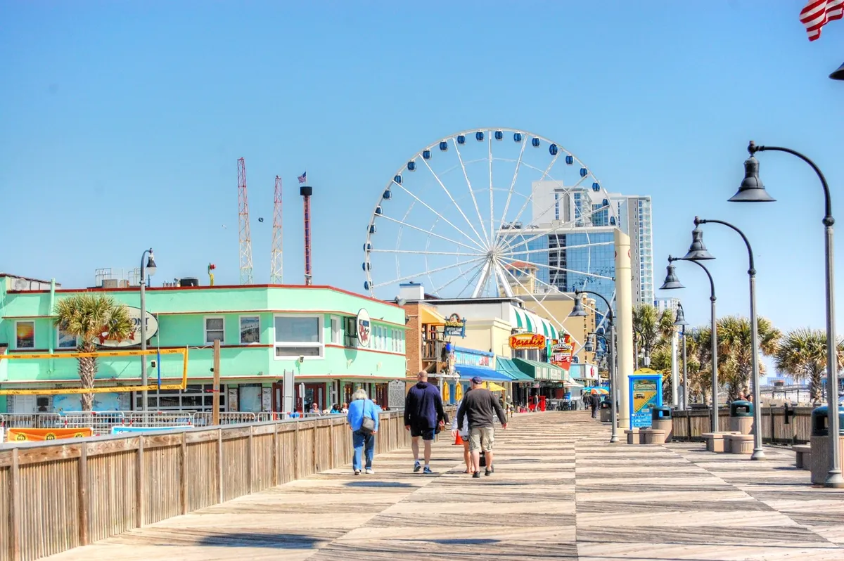 This exterior image showcases the iconic Myrtle Beach boardwalk, with a prominent ferris wheel, vibrant colorful buildings, and people enjoying a stroll. The scene is lively and filled with activity, capturing the joyful and inviting atmosphere of Myrtle Beach's famous attractions.