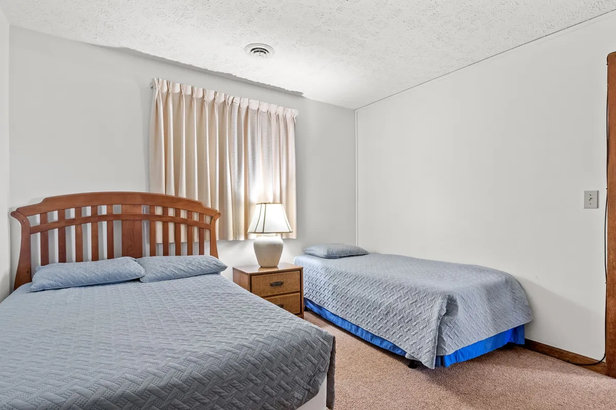 A bedroom with a wooden headboard Queen or Full bed and an additional Twin bed, with a lamp on the nightstand and curtains framing the window. The atmosphere is comfortable and welcoming, ideal for families or friends sharing the space.