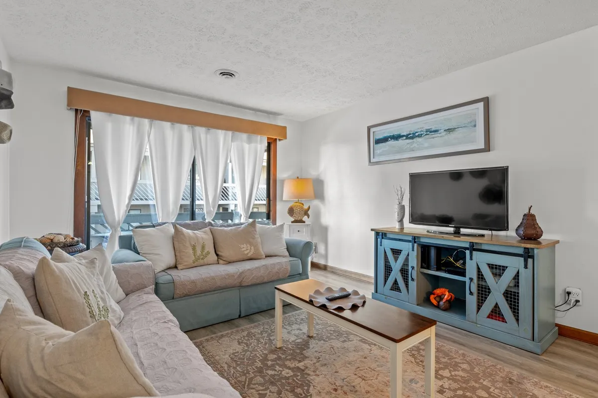 The image showcases a living area with a plush sofa, inviting coffee table, entertainment-ready TV stand, and soft curtains filtering in natural light, all accented by a warm lamp. The space is arranged for relaxation, conversation, and cozy family evenings, setting a welcoming tone for the entire condo.