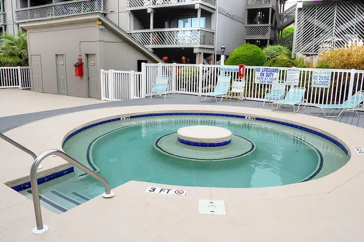A sun-drenched pool area featuring a large hot tub, several lounge chairs, and lush landscaping. The inviting space showcases an amenity-rich setting, ideal for relaxing, socializing, and soaking up the sun, mirroring the atmosphere of a well-appointed resort complex.