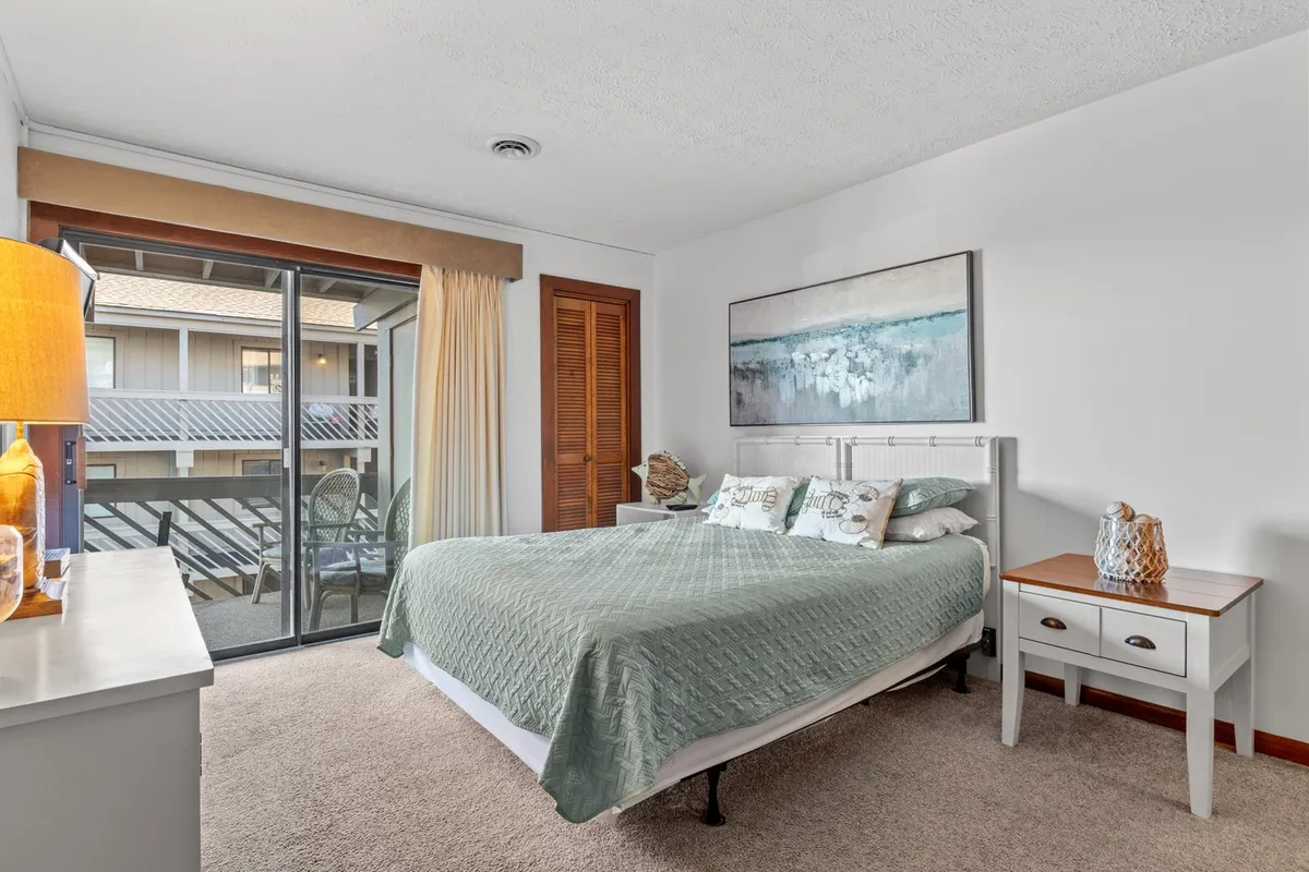 This image shows a serene bedroom featuring a bed with a green quilt, a sliding glass door letting in natural light, and abstract artwork on the wall. The overall feel is tranquil and restful, with soft linens and uncluttered décor.