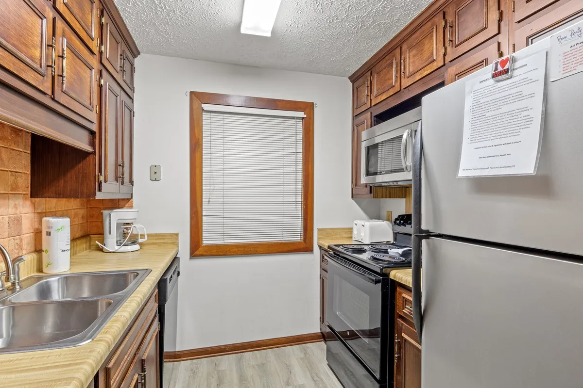 The image showcases a warm, inviting kitchen featuring modern appliances, wood cabinetry, and a window with blinds that allows natural light to fill the space. The countertop provides ample workspace for meal prep, and the layout encourages gathering and conversation.