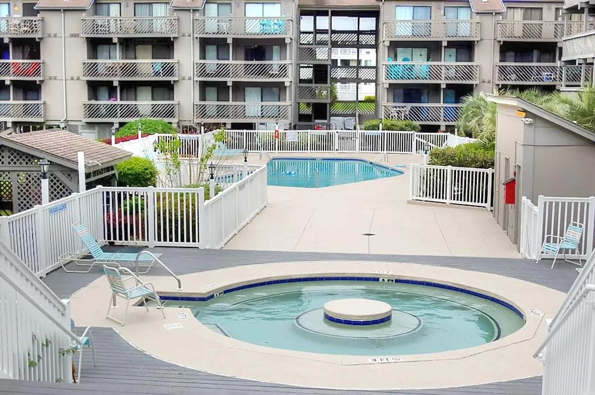 A hot tub and swimming pool sit in the midst of surrounding buildings, with lounge chairs set up around the water for sunbathing or socializing. The tranquil setting suggests both lively group activities and quiet relaxation moments.