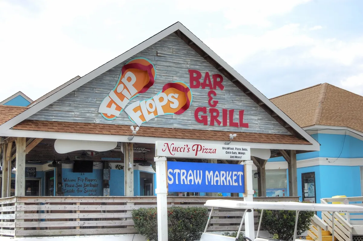 A vibrant exterior shot displaying the signage for Flip Flops Bar & Grill, Nucci’s Pizza, and the Straw Market, with outdoor dining and shopping spaces that suggest a fun, communal environment.